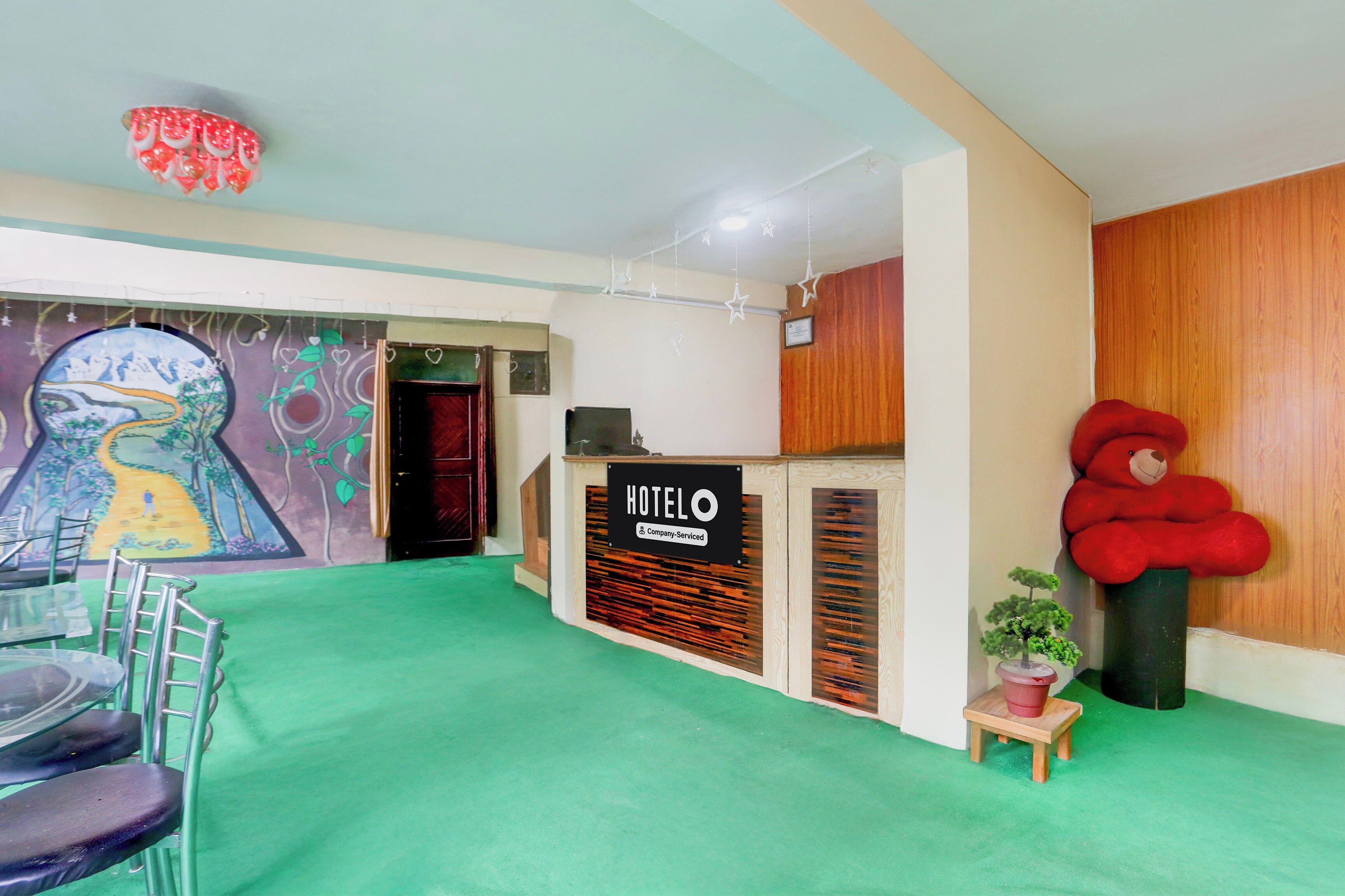 A hotel reception area with a green carpet, a reception desk, a wall-mounted teddy bear, and a mural with a landscape.