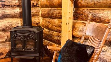 A log cabin interior with a black wood stove, a wooden rocking chair with a black cushion, and a white heater.