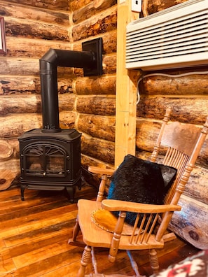 A log cabin interior with a black wood stove, a wooden rocking chair with a black cushion, and a white heater.