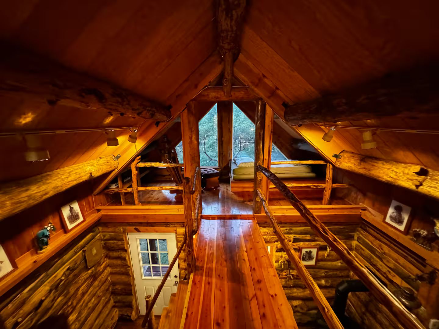 A log cabin interior with a central wooden staircase, a door, and a window providing a view of the outdoors.