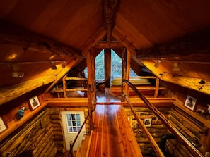 A log cabin interior with a central wooden staircase, a door, and a window providing a view of the outdoors.