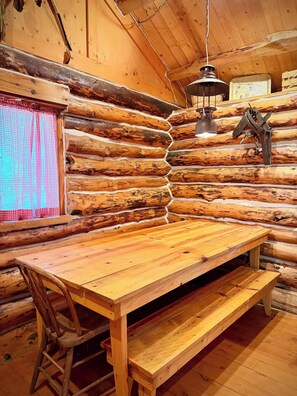 A log cabin interior with a wooden table and bench, a hanging lantern, and a window with a checkered curtain.