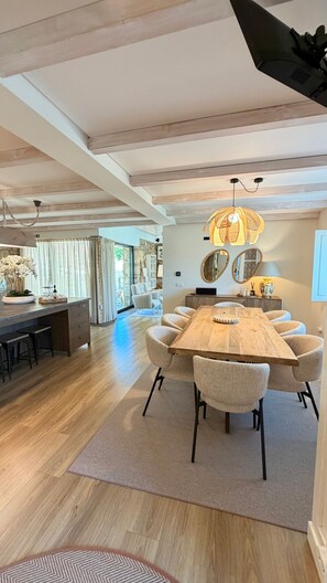 Ampla área de refeições com uma grande mesa de madeira e seis cadeiras, um candelabro e vista para a sala de estar adjacente.