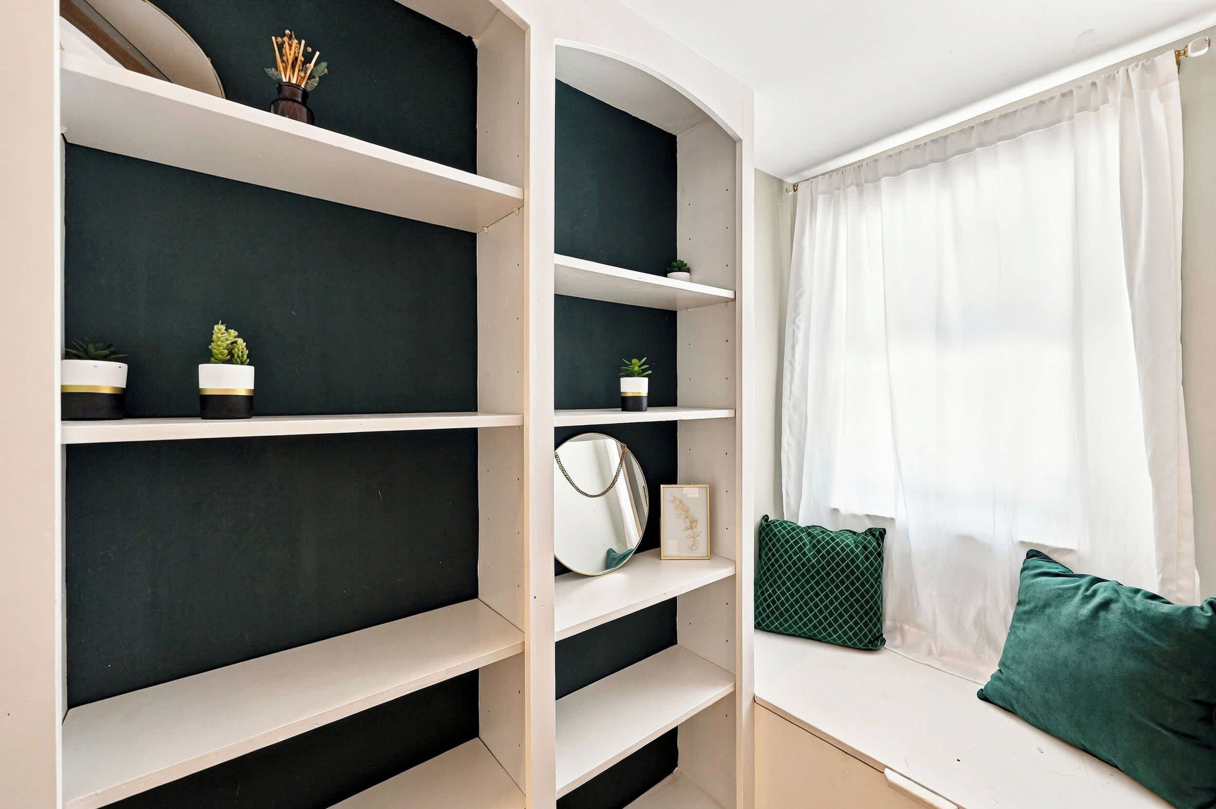 Una habitación con estantería empotrada, ventana con cortinas traslúcidas y un cojín verde en un banco.