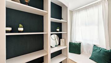 Una habitación con estantería empotrada, ventana con cortinas traslúcidas y un cojín verde en un banco.