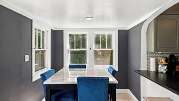 Un comedor con una mesa de mármol y sillas tapizadas en azul, ventanas amplias y una cocina con encimera negra.
