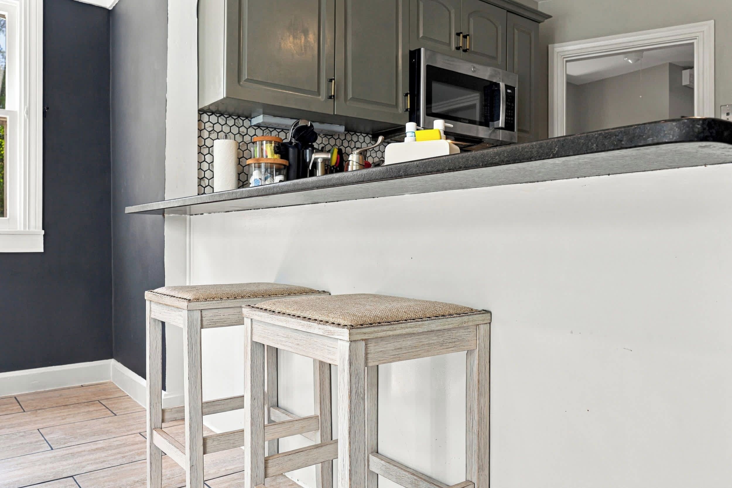Una cocina con gabinetes grises, una encimera negra y dos taburetes de madera con asientos trenzados.