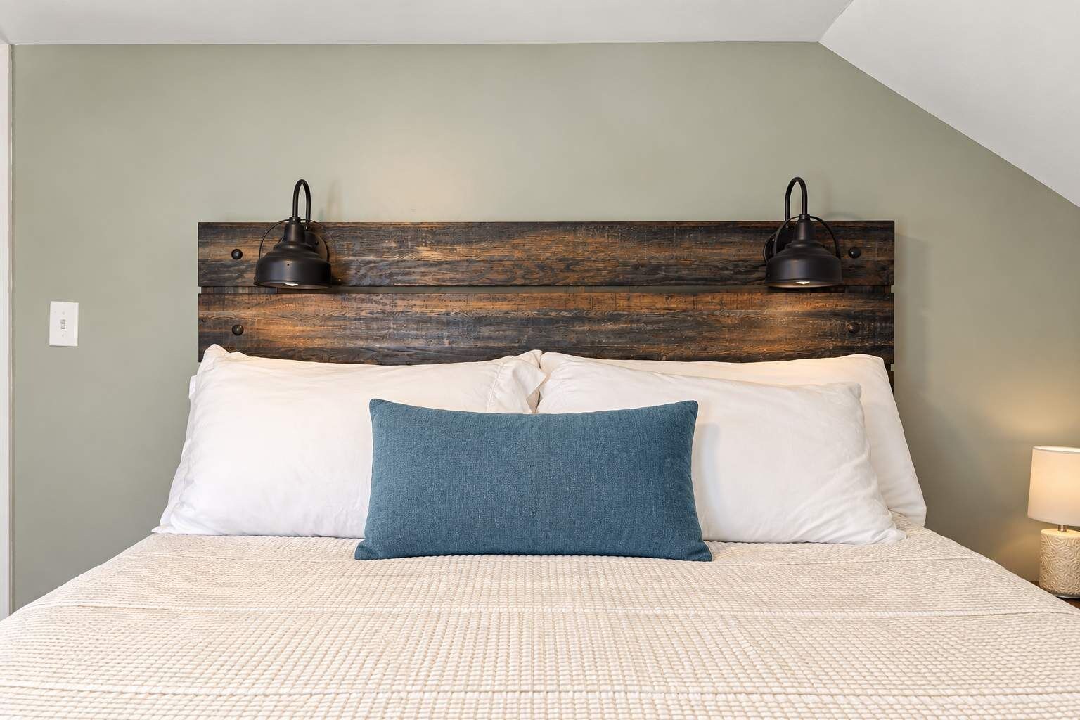 Un lit recouvert d’une couverture blanche texturée, deux oreillers blancs, un coussin décoratif de couleur turquoise et une tête de lit en bois agrémentée de deux lampes fixées au mur.