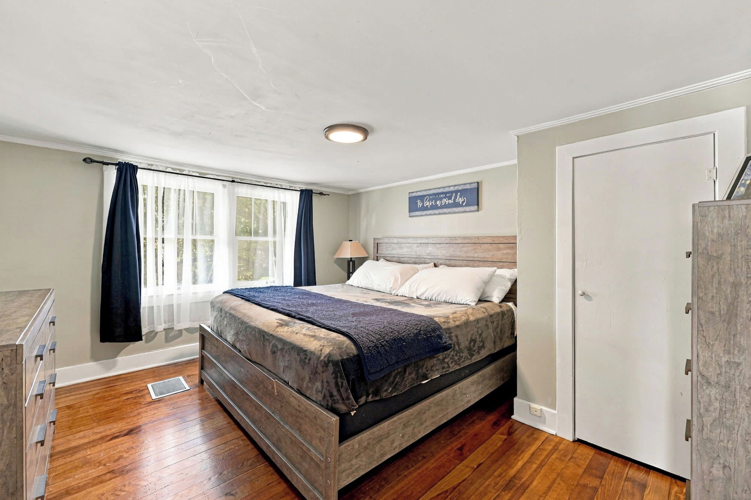 Un dormitorio con cama, mesita de noche, lámpara, ventana con cortinas y puerta.