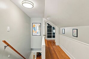 A hallway with a wooden staircase, a framed picture of flowers, and a door leading to a room with a white door and a shelf with decorative items.