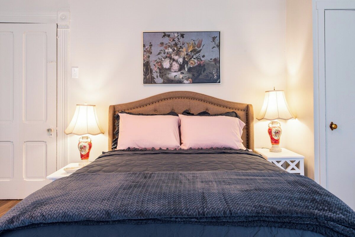 A bedroom with a bed, two bedside lamps, a wall-mounted painting, and a white door.