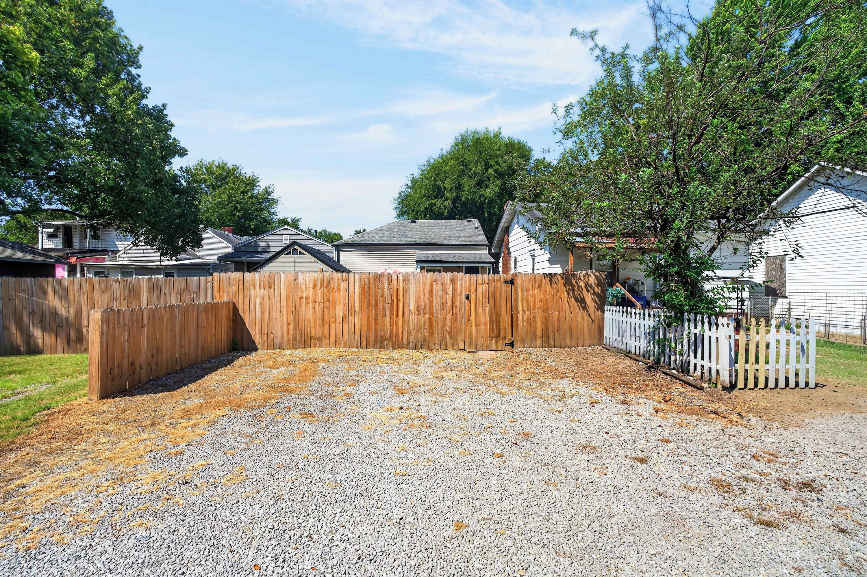 A residential backyard with a wooden fence, gravel ground, and houses in the background.