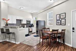 A modern kitchen with white cabinets, a central island with bar stools, a dining table with chairs, and a refrigerator.