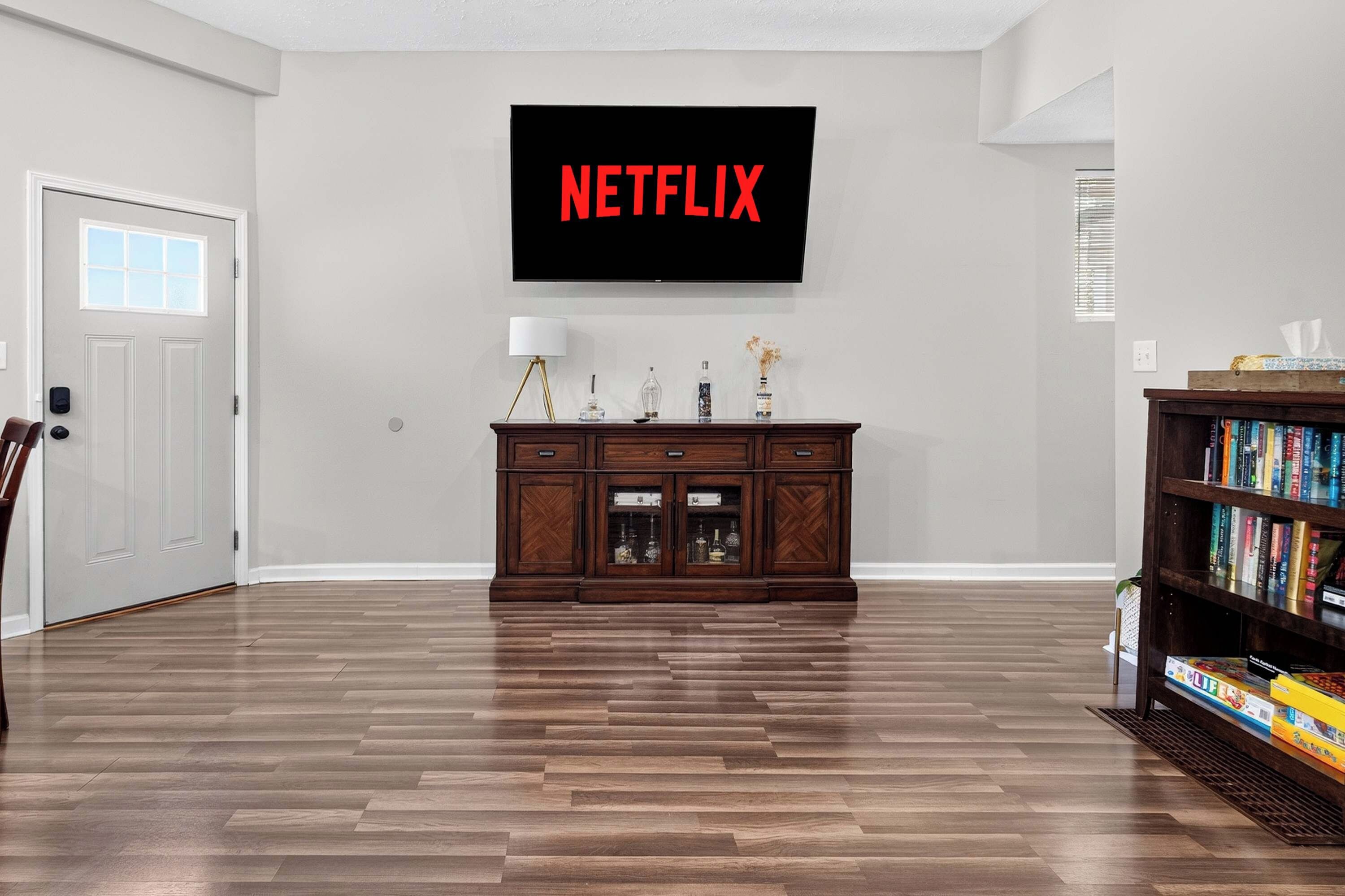 A living room with a flat-screen TV mounted on the wall displaying 'NETFLIX', a wooden cabinet with decorative items, and a bookshelf filled with books.