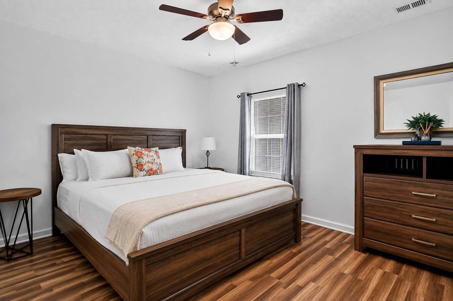 A bedroom with a wooden bed, a nightstand, a dresser, a ceiling fan, and a window with curtains.
