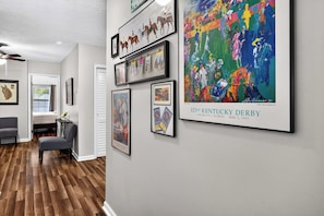 A hallway with framed artwork, a bed, a chair, and a table.