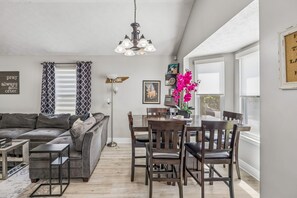 Una sala de estar moderna con un sofá gris, una mesa de comedor con sillas, una araña y un ventanal con vistas.