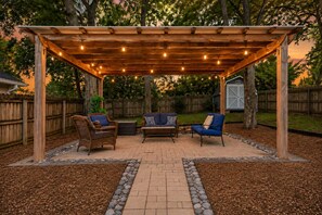 Un patio al aire libre con una pérgola, dos sillas, una mesa y una guirnalda decorativa de luces.