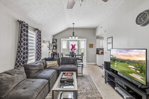 A living room with a gray sectional sofa, a coffee table with books, a television on a stand, and a view of a golf course. - Iconic Music City retreat w/ backyard and hot tub! (Nashville)