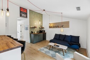 Ein modernes Wohnzimmer mit einem dunkelblauen Sofa, einem Holztisch und einer Küchenzeile mit grüner Spritzschutz und einem hängenden Schild.