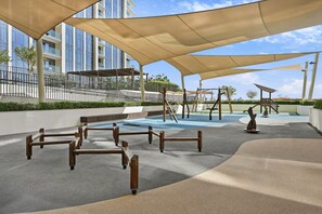 An outdoor play area with swings and a climbing structure, surrounded by shaded areas and a building in the background. - Premium 1BR - Creek Harbour - Pool, Sea View (Dubai)