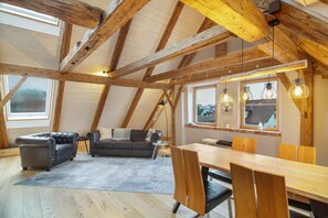 A loft-style room with exposed wooden beams, a leather sofa, a dining table with chairs, and a view through a window.