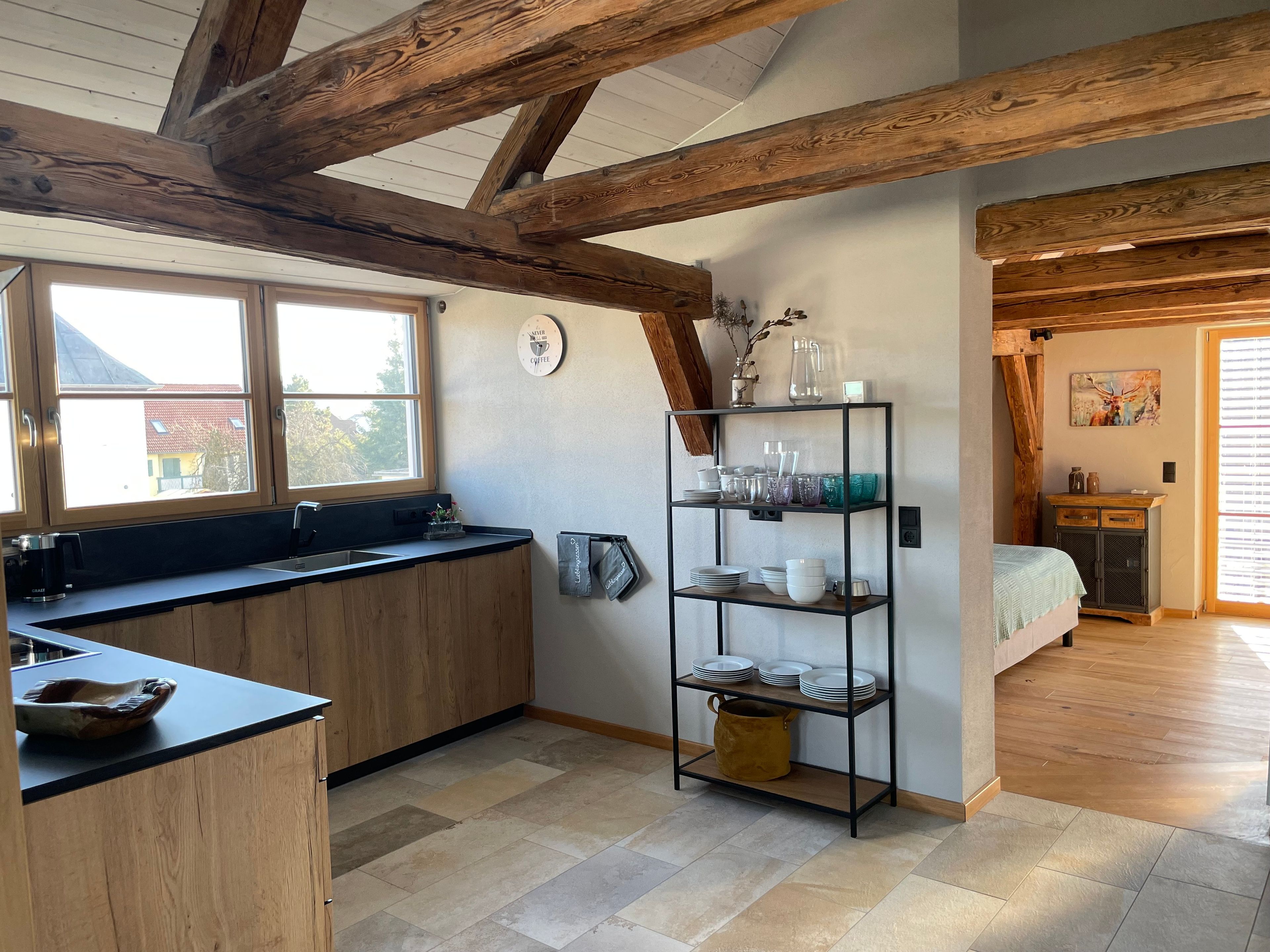 Eine Küche mit Holzbalken, einem schwarzen Metallregal mit Geschirr, einem Fenster und Blick nach draußen.
