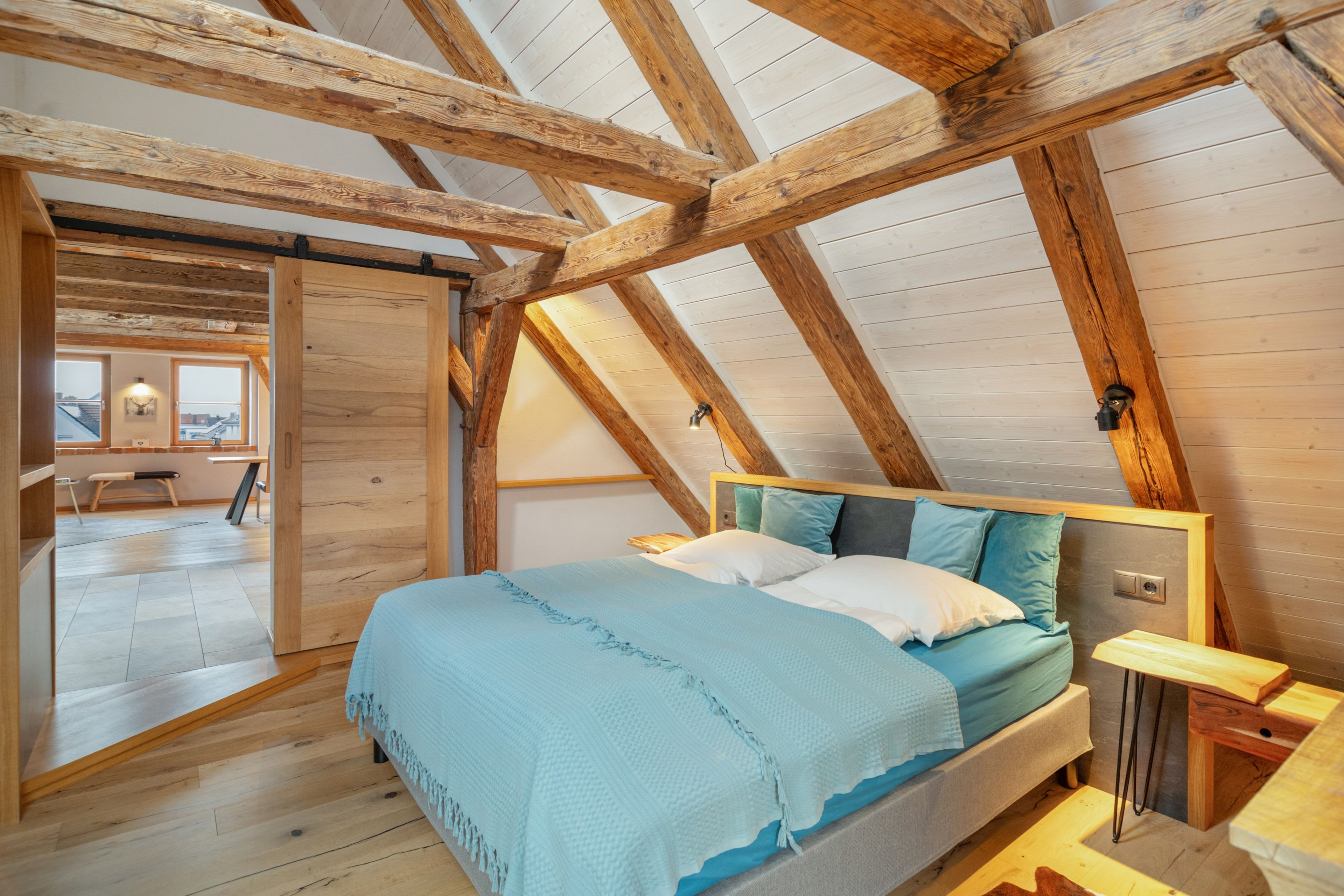 Ein Schlafzimmer mit schrägen Wänden, Holzbalken, einem Bett mit Kissen, einem Nachttisch und Blick auf die Stadt durch ein Fenster.