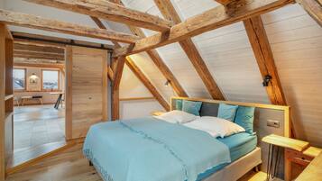 Ein Schlafzimmer mit schrägen Wänden, Holzbalken, einem Bett mit Kissen, einem Nachttisch und Blick auf die Stadt durch ein Fenster.
