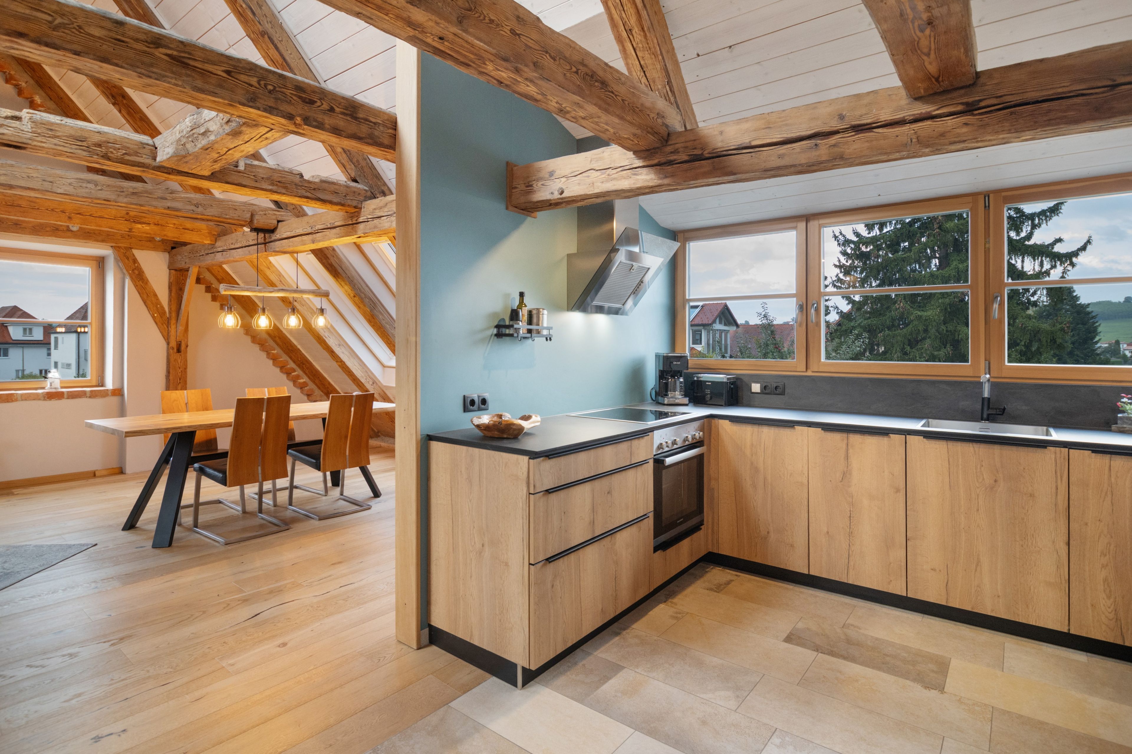Een moderne keuken met houten balken, een eethoek met tafel en stoelen, en uitzicht door grote ramen.