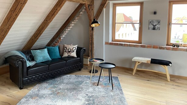 A loft-style room with exposed wooden beams, a black leather sofa, a small round table, and a bench. - DG Apartment alpenchic with Balcony in Ottobeuren – Sleeps 2 (Ottobeuren)