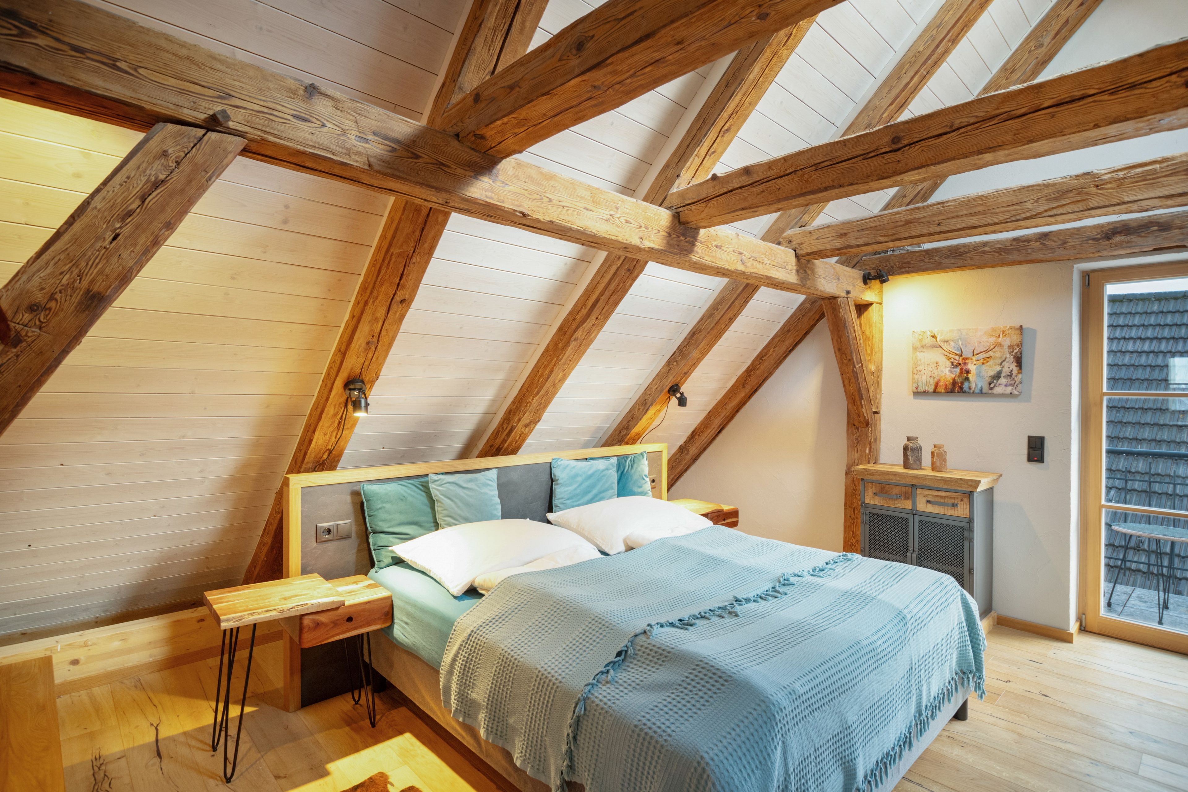 Een slaapkamer met een schuine kap, houten balken, een bed met kussens, een nachtkastje, een bureau en een raam.