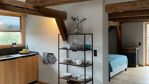 A kitchen with wooden beams, a black metal shelf with dishes, a window, and a bed in the adjacent room. - DG Apartment alpenchic with Balcony in Ottobeuren – Sleeps 2 (Ottobeuren)
