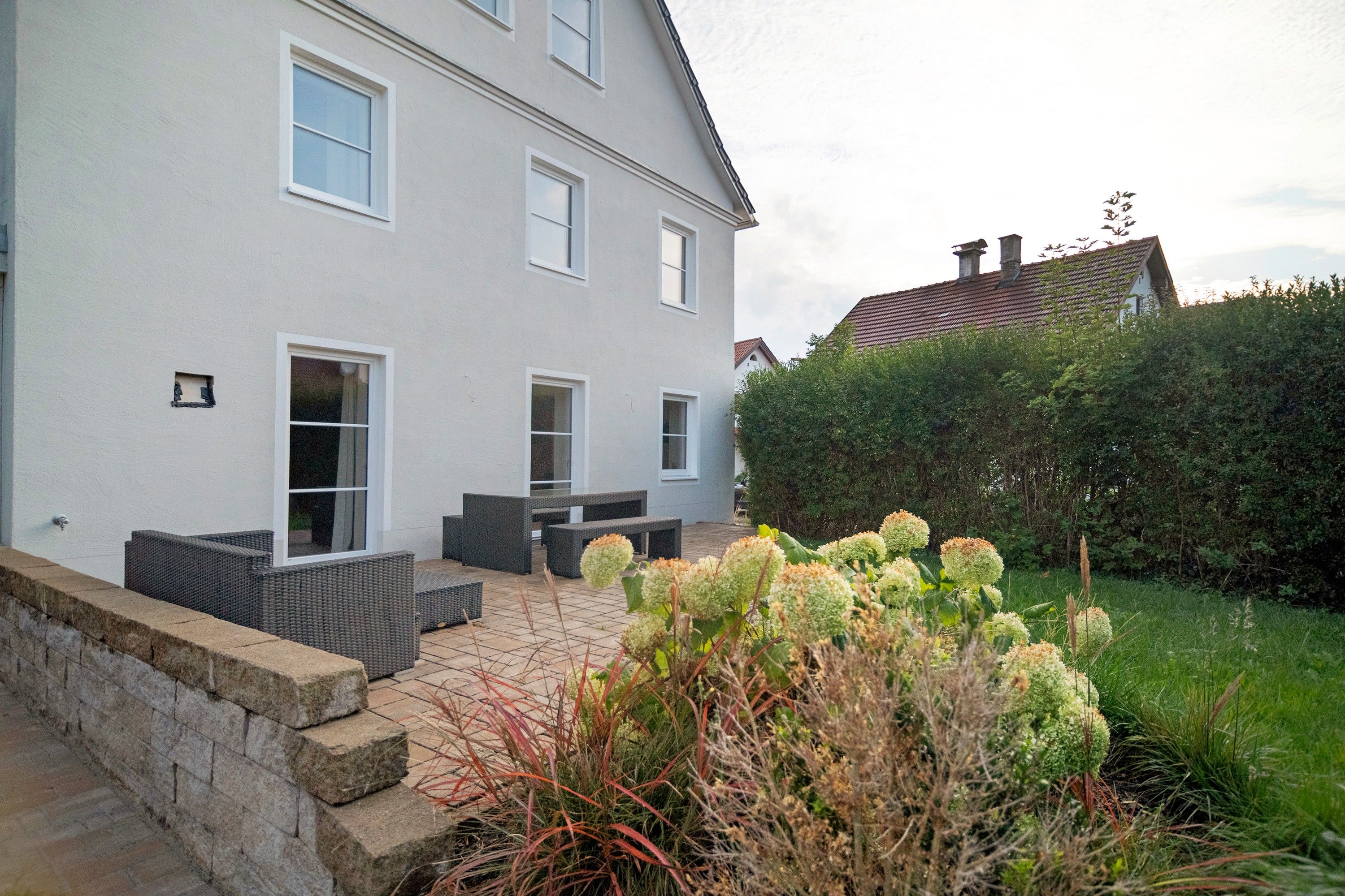 Une maison à deux étages dotée d’une terrasse dallée, d’un espace salon extérieur et d’un jardin bien entretenu.