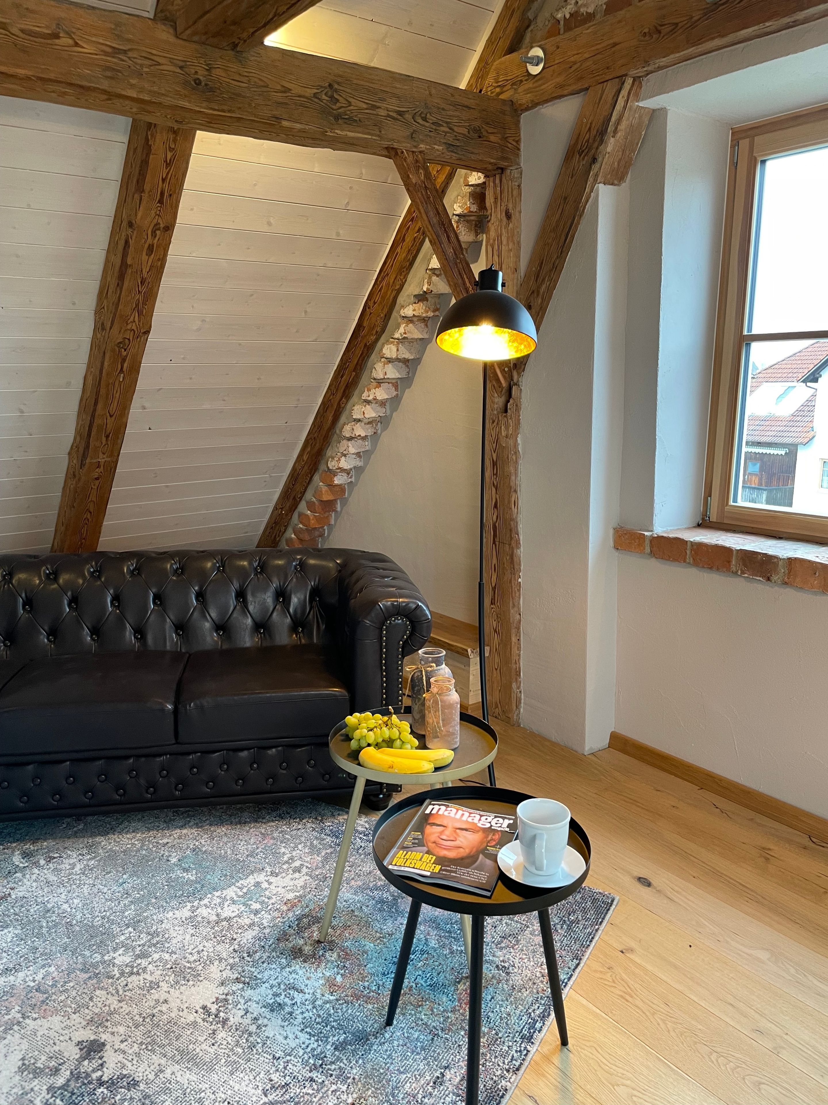 Ein Loft-Zimmer mit freiliegenden Holzbalken, einem schwarzen Ledersofa, einem kleinen runden Tisch mit einer Zeitschrift, einer Lampe und einem Fenster mit Ausblick.