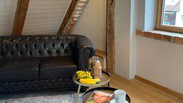Ein Loft-Zimmer mit freiliegenden Holzbalken, einem schwarzen Ledersofa, einem kleinen runden Tisch mit einer Zeitschrift, einer Lampe und einem Fenster mit Ausblick.
