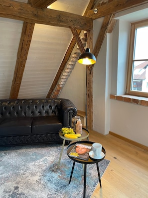 Une pièce de style « loft » avec des poutres apparentes, un canapé en cuir noir, une petite table ronde avec un magazine, une lampe et une fenêtre donnant sur un paysage.