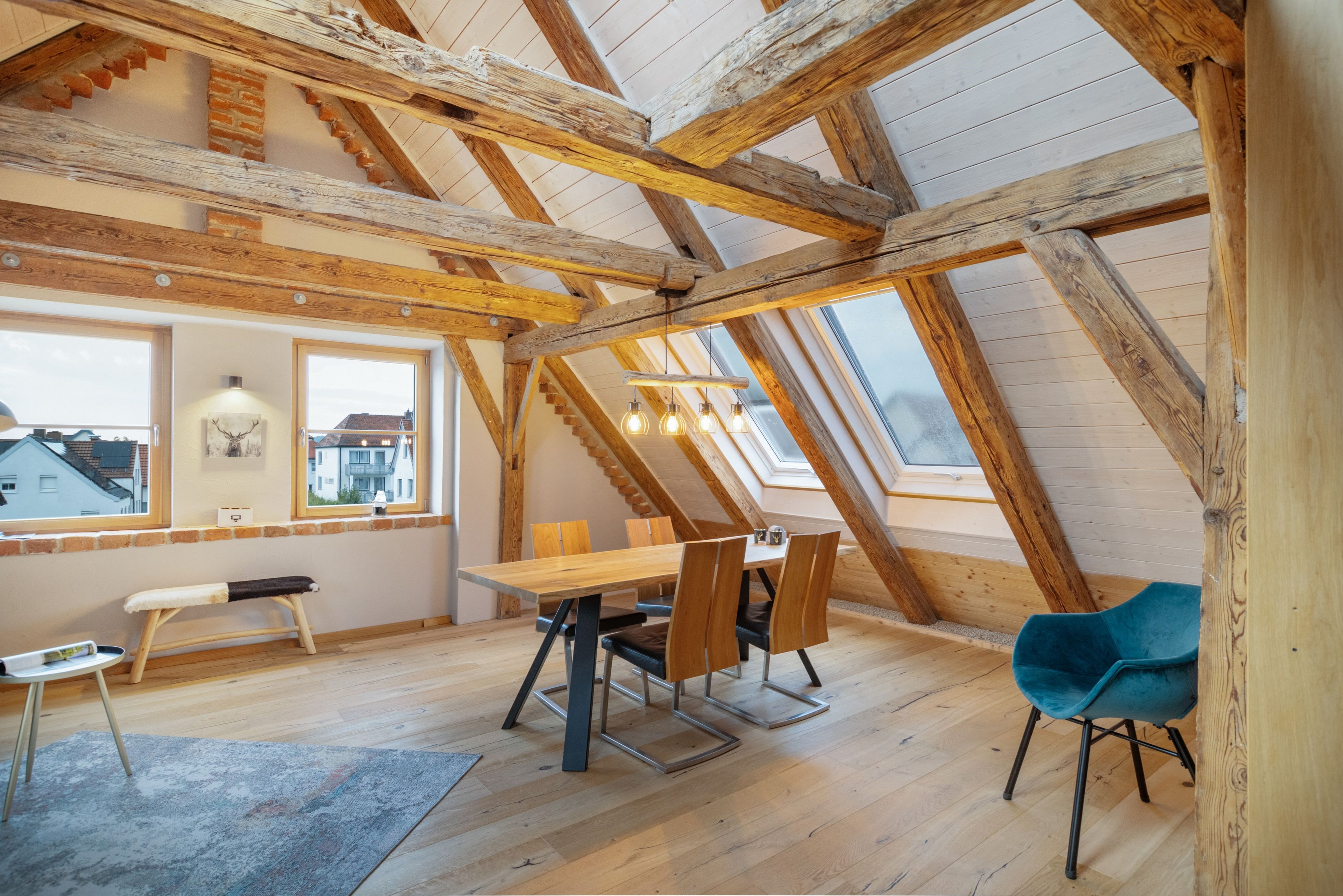 Een loft-achtige kamer met zichtbare houten balken, een eettafel met stoelen, een bankje en een uitzicht op de stad door middel van dakramen.
