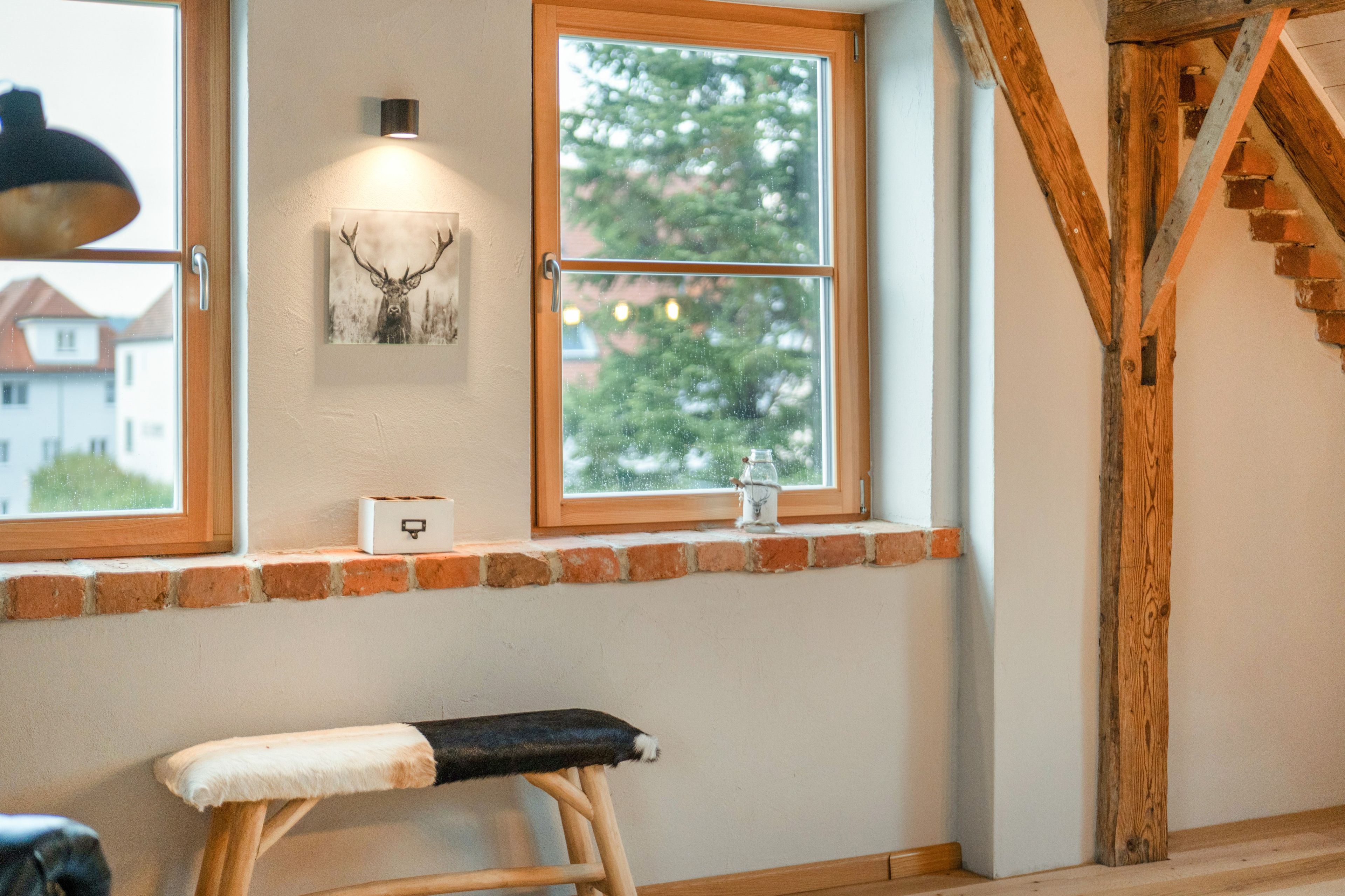 Una habitación con un banco de madera, una ventana con vistas a árboles, un cuadro colgado en la pared y una mesita con un tarro.