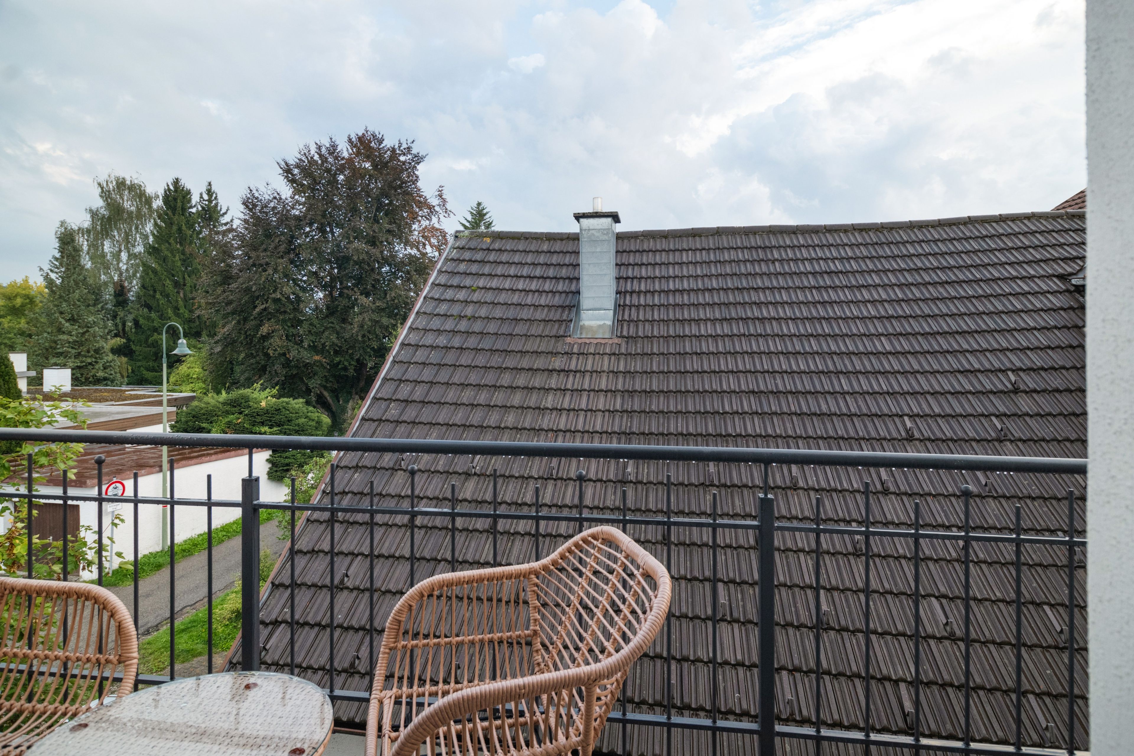 Ein Balkon mit Blick auf ein Gebäudedach, einem Metallgeländer und einem Korbstuhl.