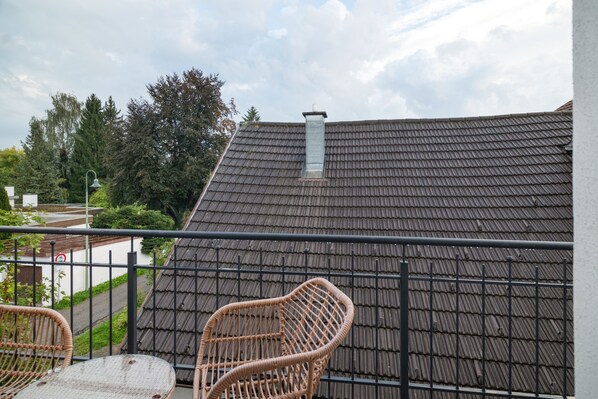 A balcony with a view of a building's roof, a metal railing, and a wicker chair.