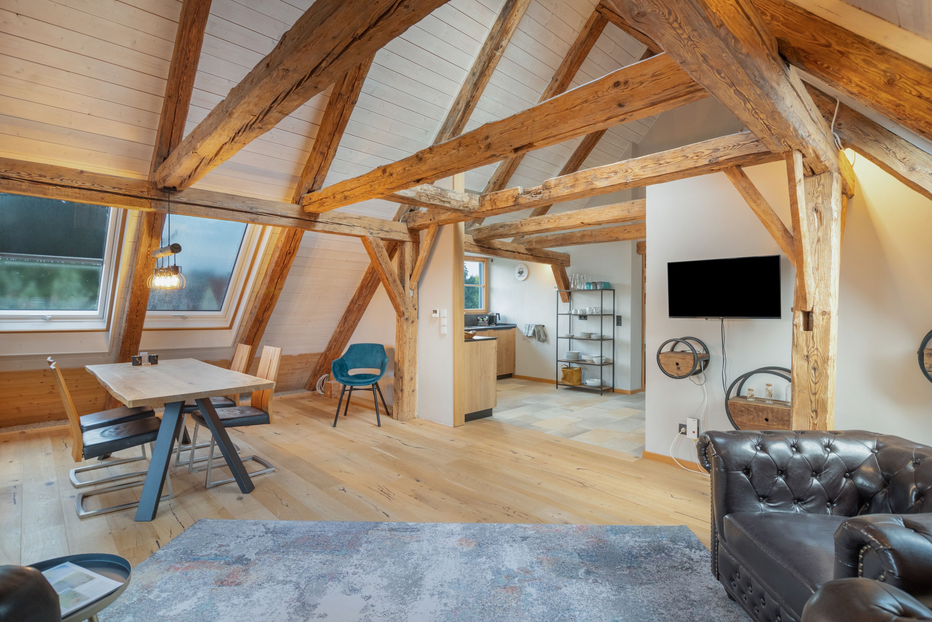 Ein Loft-Zimmer mit freiliegenden Holzbalken, einem Essbereich, einer Sitzecke mit einer Ledersofa und einer Küche im Hintergrund.