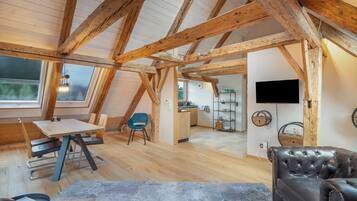 Ein Loft-Zimmer mit freiliegenden Holzbalken, einem Essbereich, einer Sitzecke mit einer Ledersofa und einer Küche im Hintergrund.