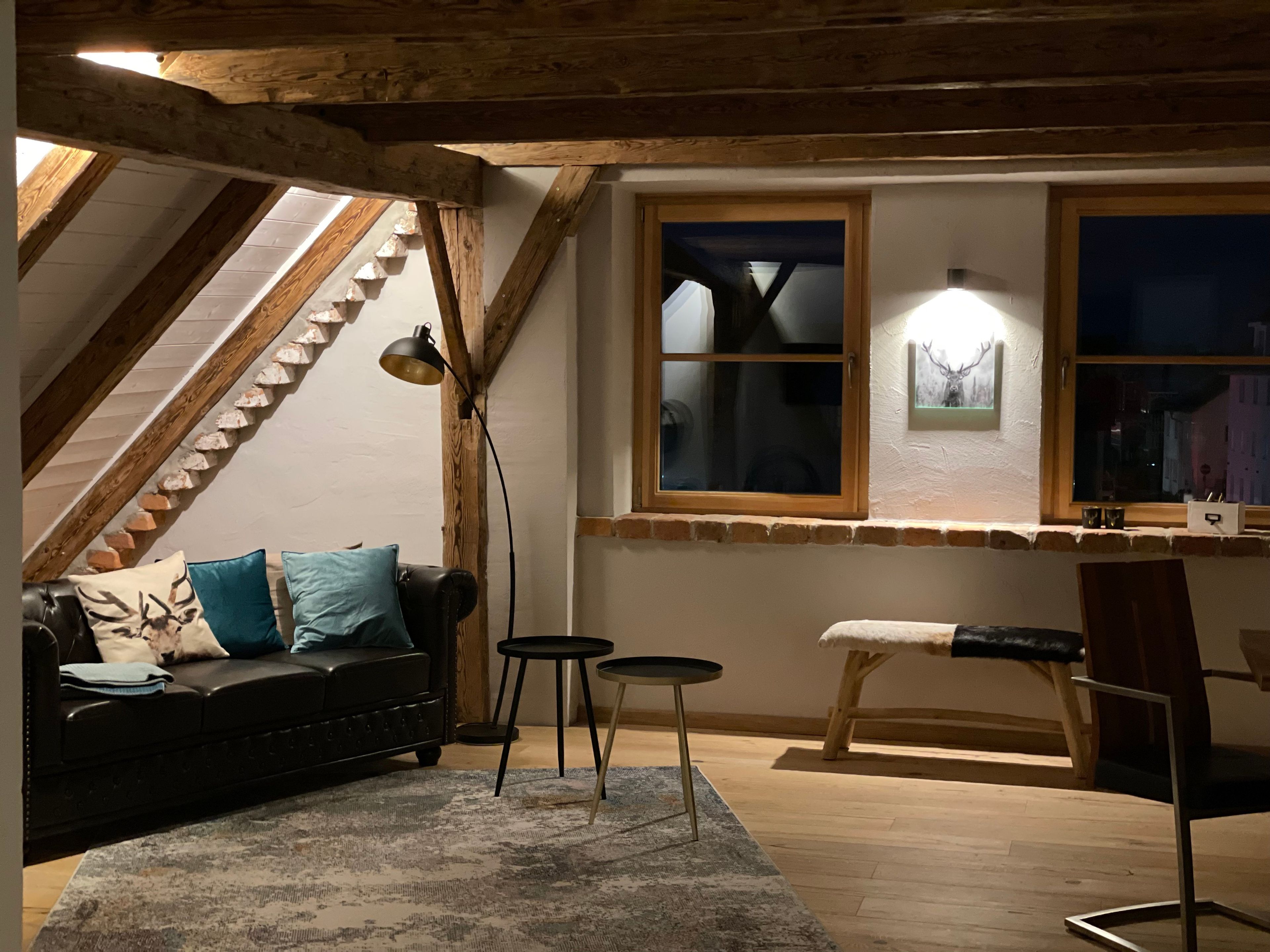Ein gem ütliches Schlafzimmer im Loft-Stil mit schrägem Dach, Holzbalken, einer schwarzen Ledersofa, einem kleinen runden Tisch und einem Holzsessel.