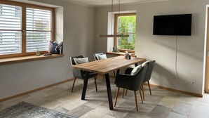 Un comedor con una mesa de madera, seis sillas, un televisor montado en la pared y una ventana con persianas.