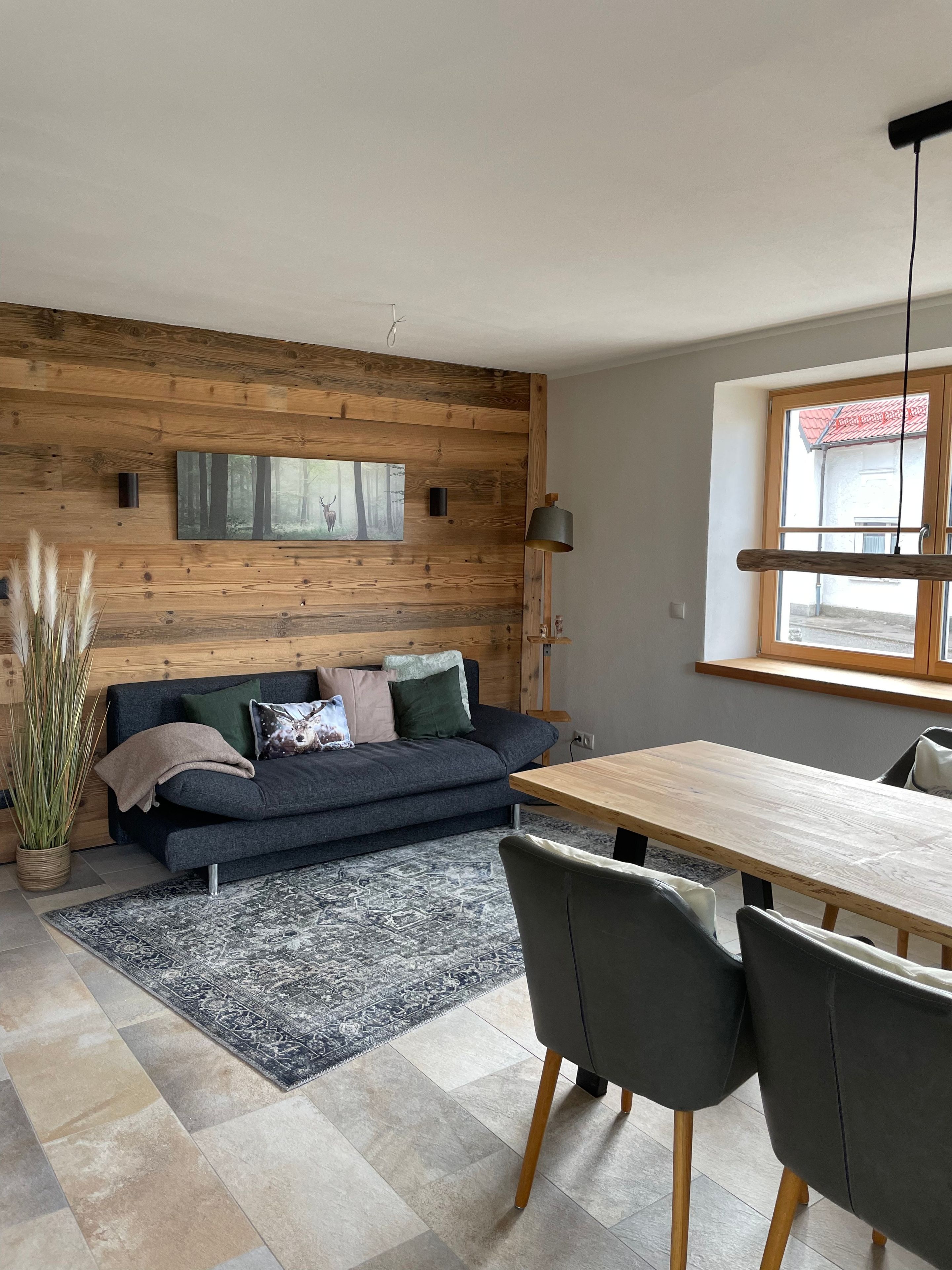 Ein modernes Wohnzimmer mit einer Holzvertäfelung, einem dunkelgrauen Sofa, einem gemusterten Teppich, einem Holzesstisch und einem Fenster mit Blick.