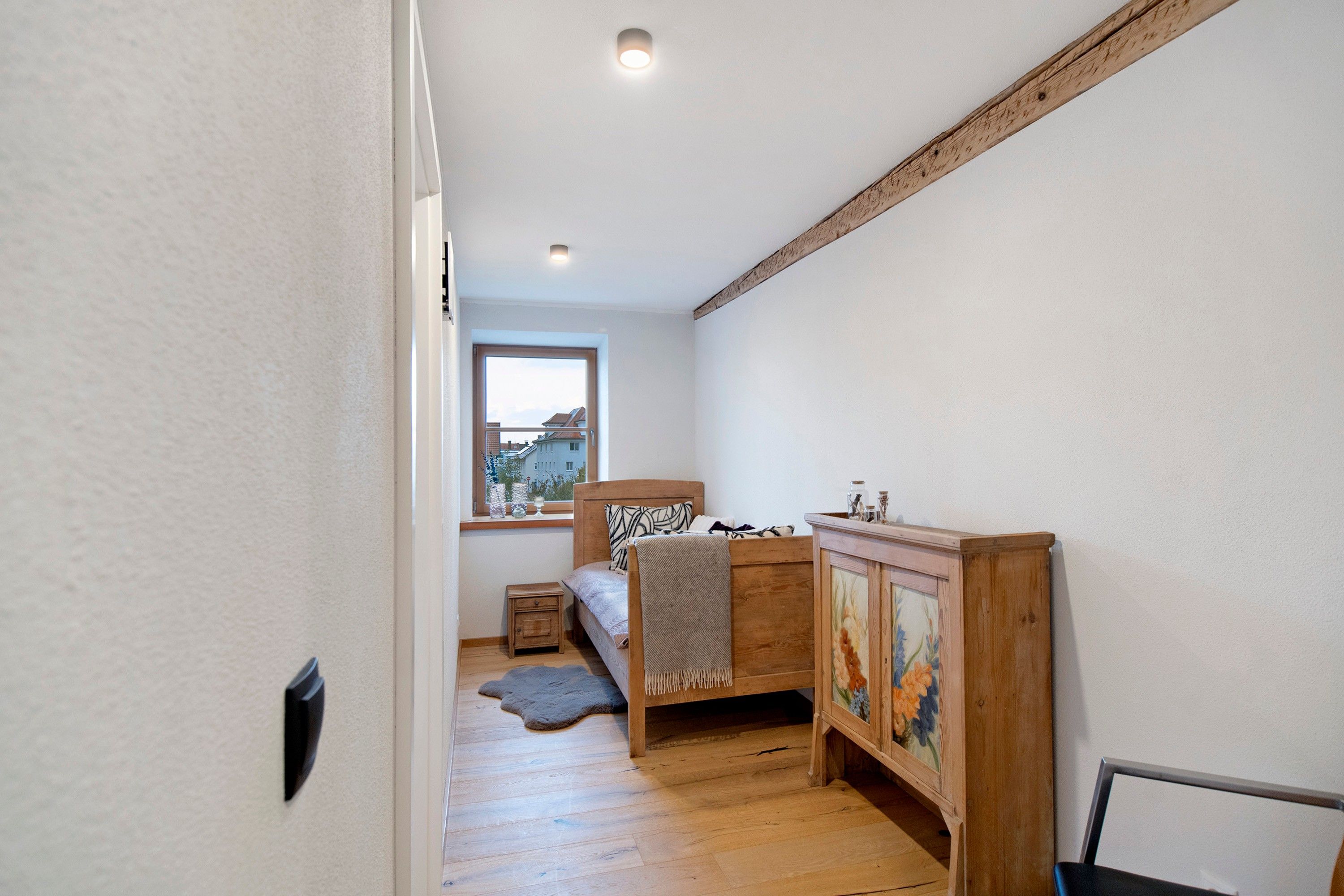 Ein enger Flur führt zu einem Schlafzimmer mit einem Bett, einem Nachttisch, einem Holzschrank mit floralem Dekor und einem Fenster mit Blick auf Gebäude.