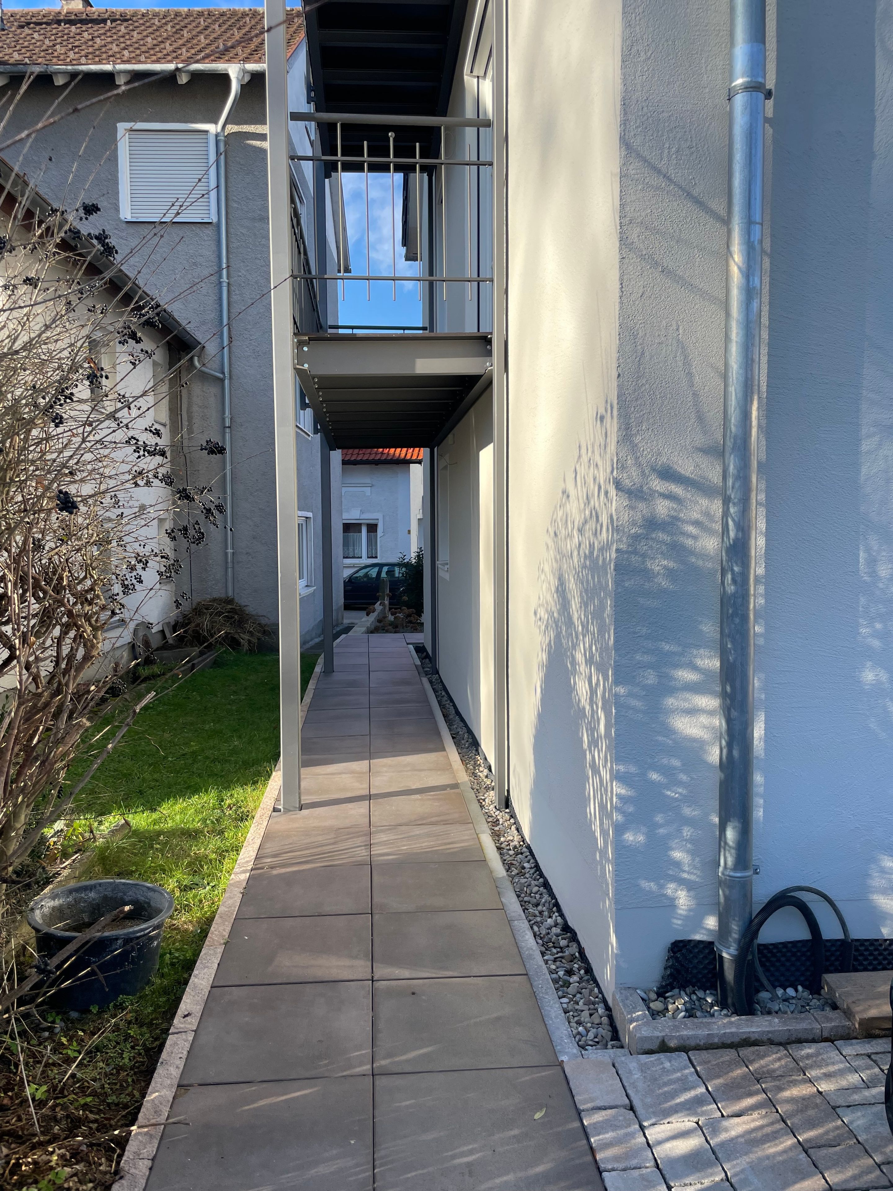 A narrow pathway leading to a building with a balcony, a tiled roof, and a clear sky.