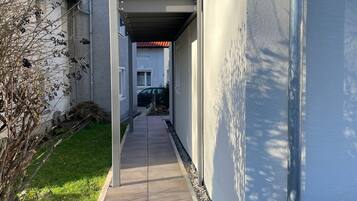 A narrow pathway leading to a building with a balcony, a tiled roof, and a clear sky.