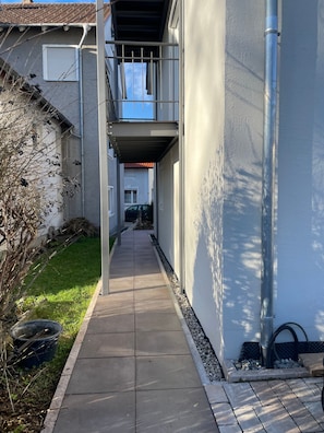 A narrow pathway leading to a building with a balcony, a tiled roof, and a clear sky.